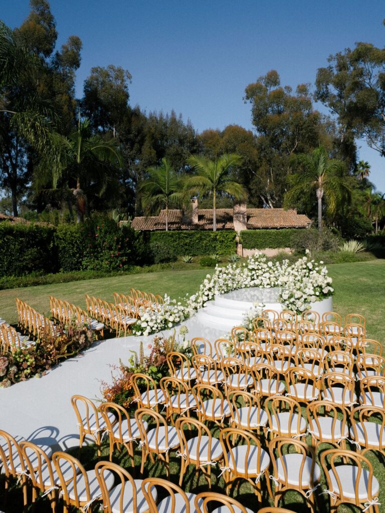 Black tie wedding ceremony on the Valencia Lawn at Rancho Valencia, with a white aisle, bentwood chairs and a half moon arch of white flowers