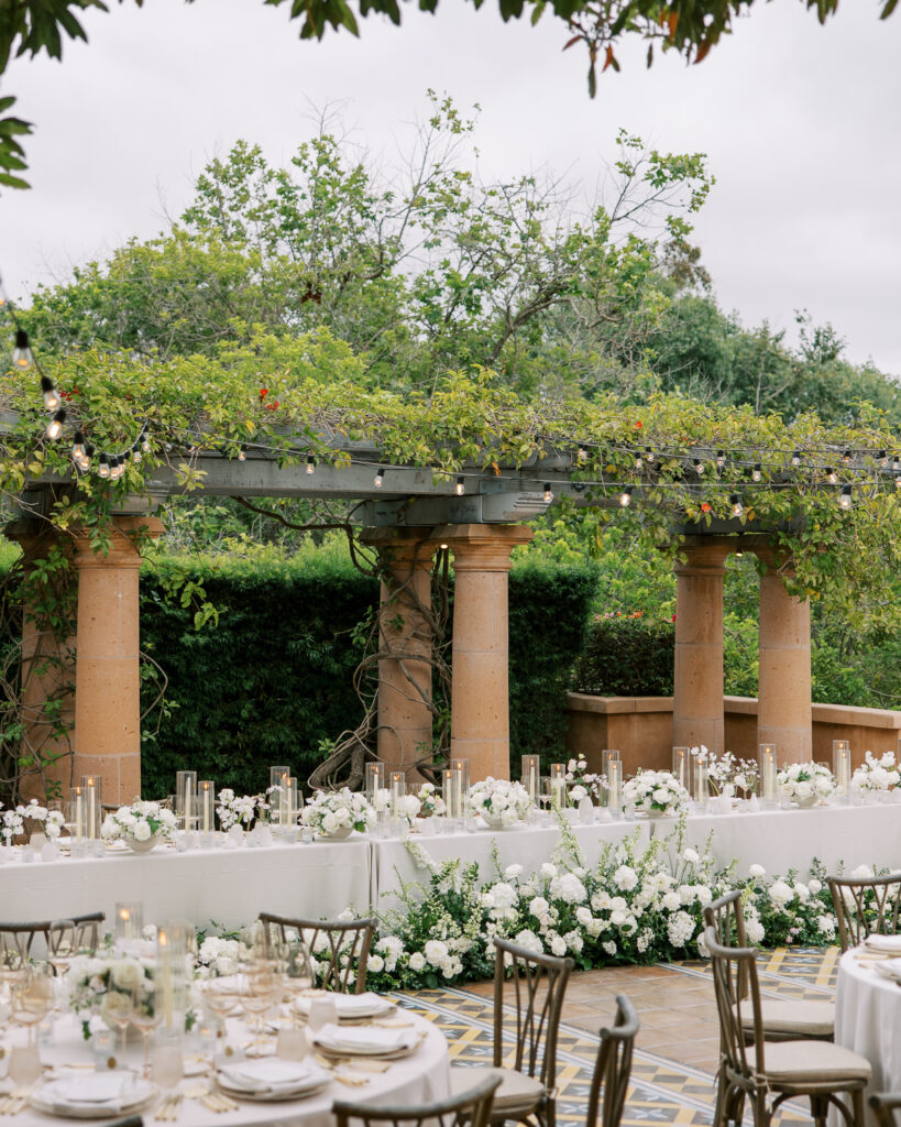 Long reception tables with white florals beneath a garden pergola, prepared from a wedding production schedule