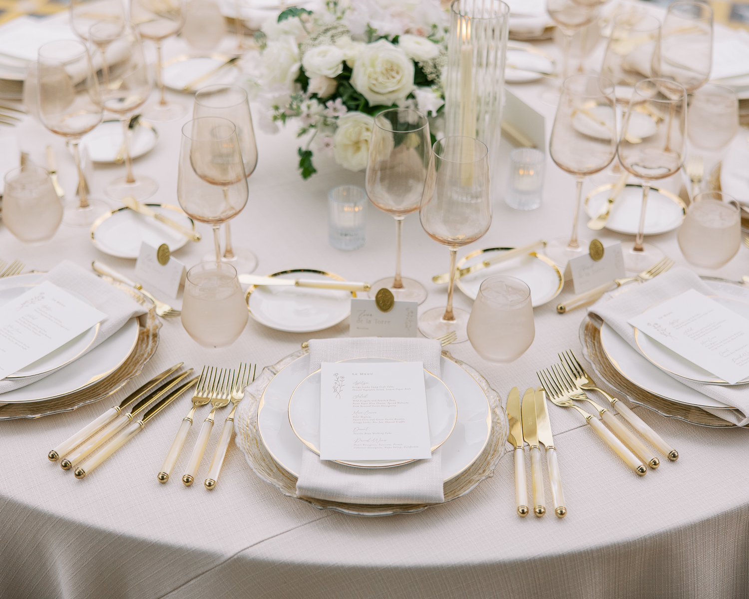 Round reception table with gold flatware, menu cards and white florals, set from a wedding weekend production schedule