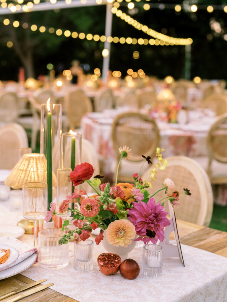 Candlelit centerpiece of lush dahlias and ranunculus on a wedding reception table under string lights