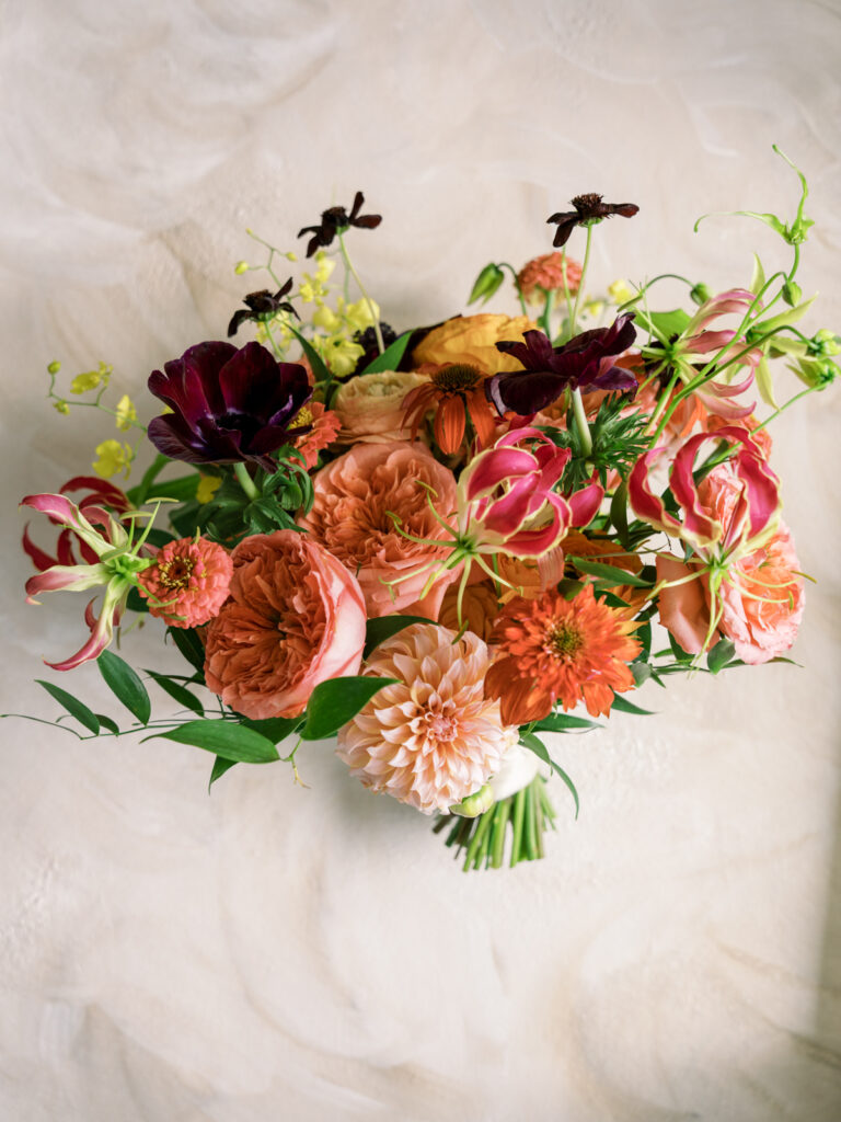 Lush bridal bouquet of coral, burgundy and blush blooms resting on a plaster surface